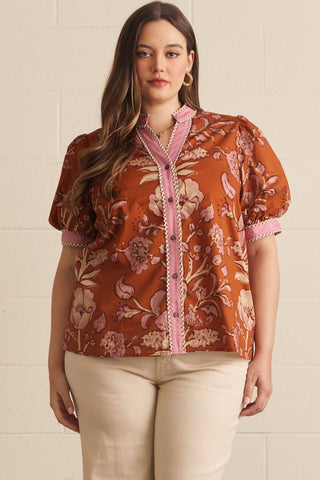 A woman modeling a rust-orange short-sleeve button-up blouse with a pink floral paisley print and pink trim, paired with beige pants, standing against a light beige brick wall.