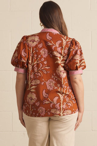 A woman shown from the back wearing a rust-orange short-sleeve blouse with a pink and beige floral pattern and beige pants, standing against a light beige brick wall.