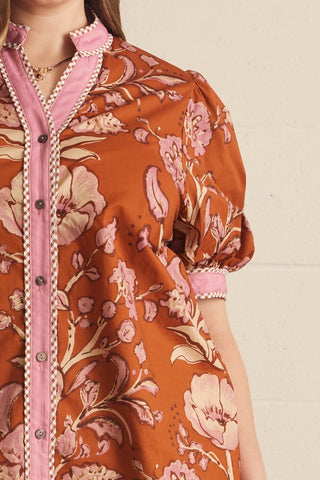Close-up of a short-sleeve button-front blouse with a rust-orange floral print and pink trim, worn by a model against a light neutral wall.