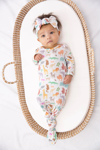 A baby lying on a white padded mat inside a woven oval bassinet, wearing a long-sleeve footed sleeper with a colorful safari animal print and a matching bow headband.