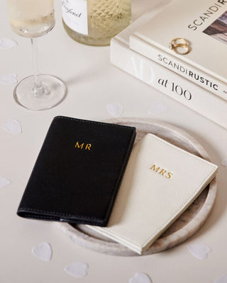 Black 'MR' and beige 'MRS' wallets on a table with books and glasses.