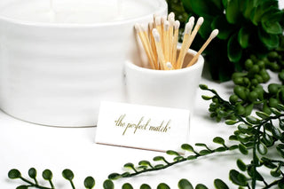 A white ceramic candle holder with a white wax candle and a small white cup holding wooden matchsticks, with a small white card reading "the perfect match" in gold script. Green foliage decor surrounds the items on a white surface.